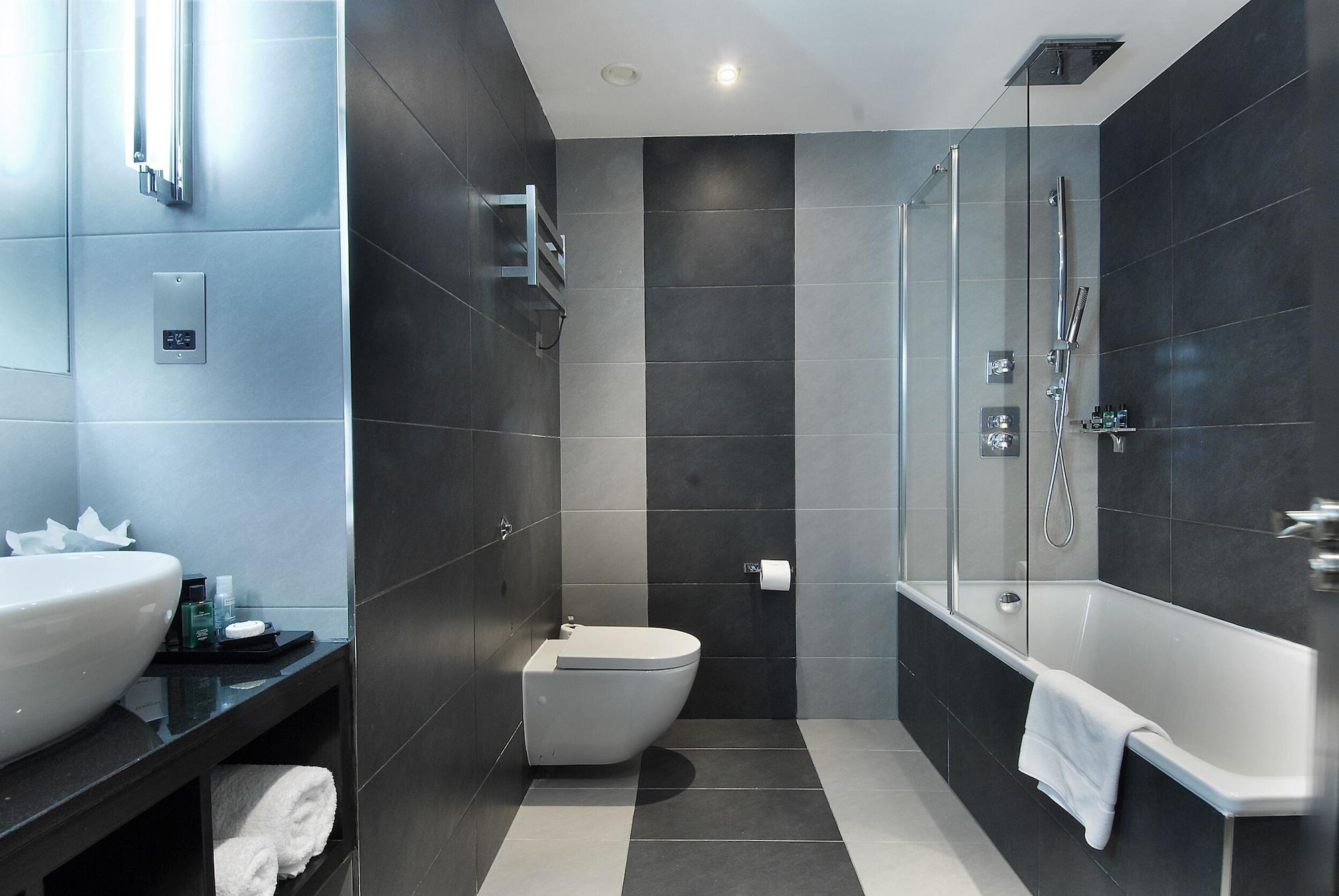 Montcalm Brewery Townhouse Hotel London Ngoại thất bức ảnh The photo shows a modern bathroom featuring a sleek design. The walls are tiled in a combination of dark gray and light gray, creating a contemporary look. On the left, there is a countertop with a basin and some toiletries placed on it. The middle s