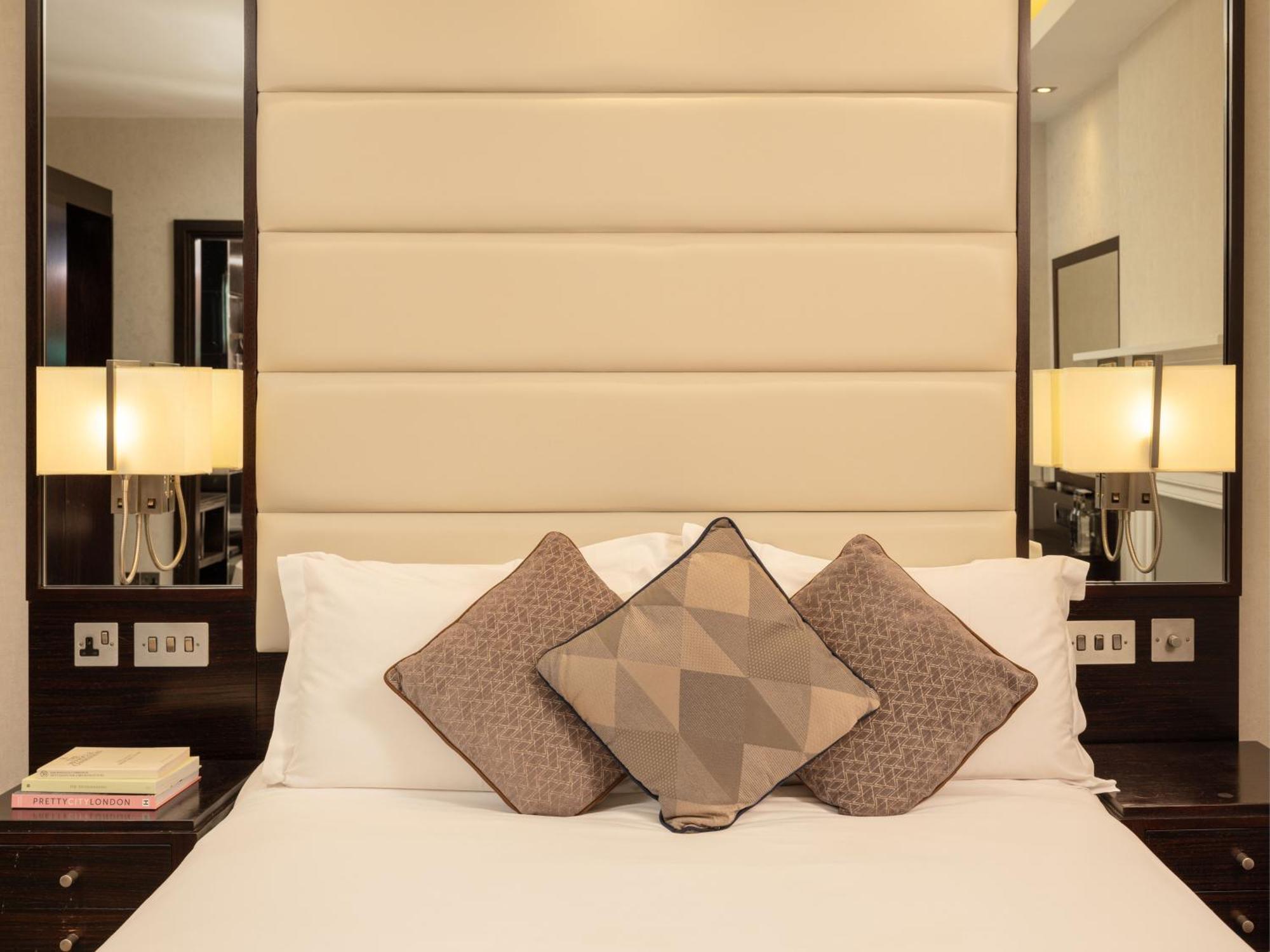 Montcalm Brewery Townhouse Hotel London Ngoại thất bức ảnh The image shows a cozy bedroom setting. The bed features a headboard upholstered in a light beige fabric. It is adorned with three decorative pillows in varying patterns and colors, placed on white bedding. On either side of the bed, there are modern