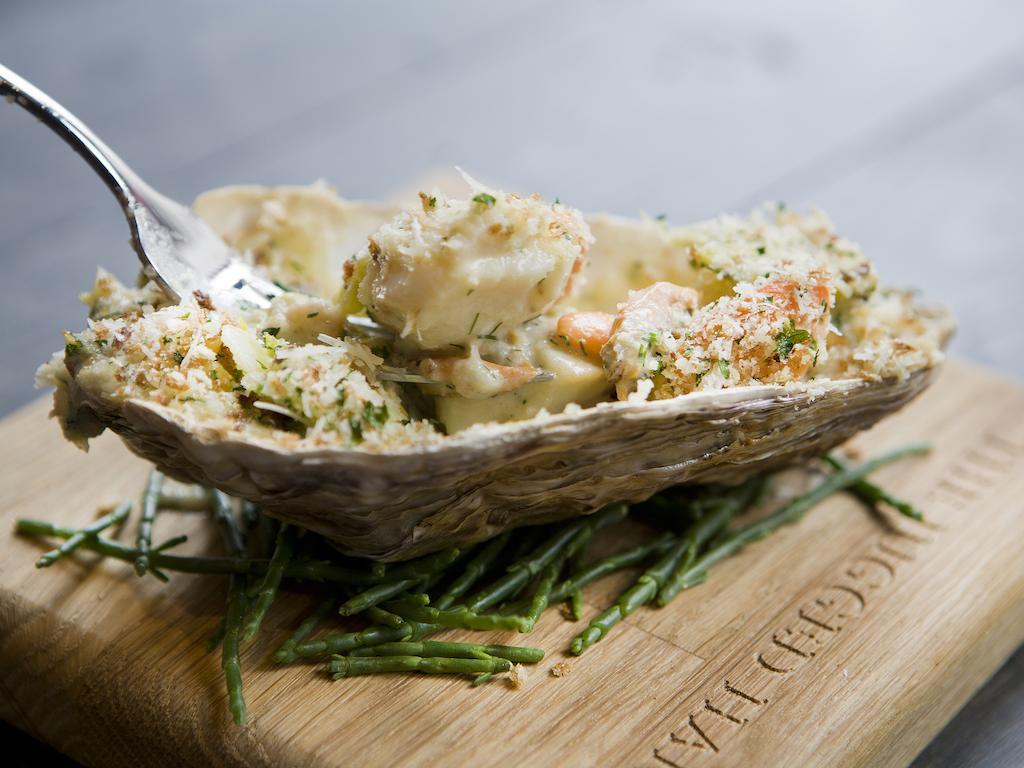 Montcalm Brewery Townhouse Hotel London Ngoại thất bức ảnh The photo shows a large oyster shell filled with a mixture that appears to include seafood, herbs, and breadcrumbs. The dish is presented on a wooden board with some greenery, possibly a type of seaweed or garnishing. A fork is placed beside the oyst